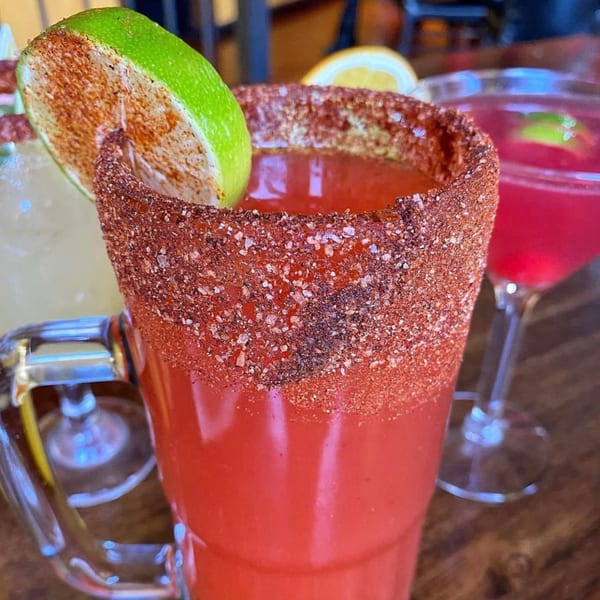 A Sonrisa Grill michelada with a chili-salt rim and lime wedge, with other drinks blurred in the background.