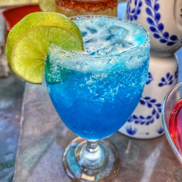 A vibrant blue margarita with a salted rim and lime wedge, next to a decorative ceramic pitcher.