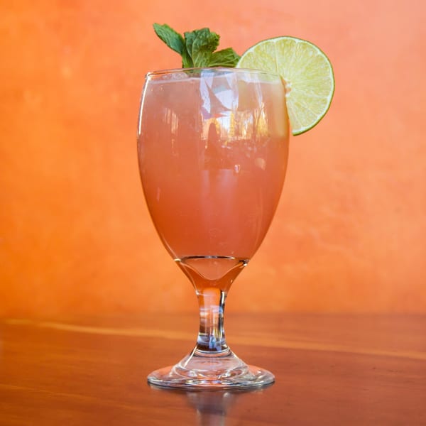 A pink Sonrisa Grill cocktail garnished with a lime slice and mint, against an orange wall.