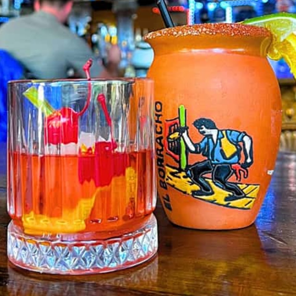 Two Sonrisa Grill cocktails, one in a glass with cherries, the other in an orange mug.