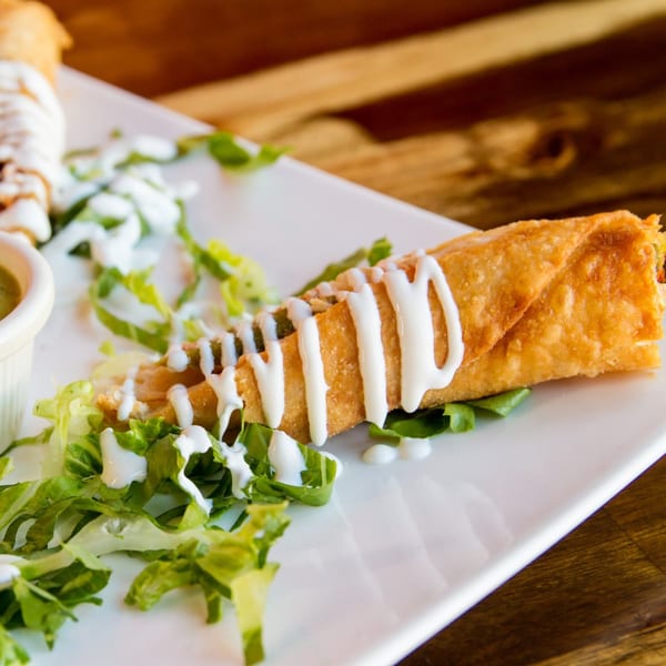 Crispy Sonrisa Grill taquito drizzled with crema, served on a bed of lettuce with a side of green sauce.