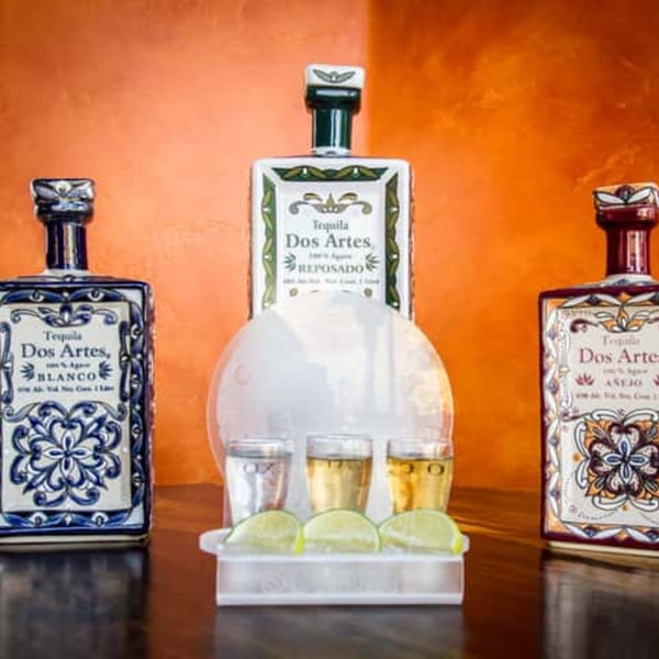 Three decorative bottles of Dos Artes tequila with a flight of three tequila shots and lime wedges.